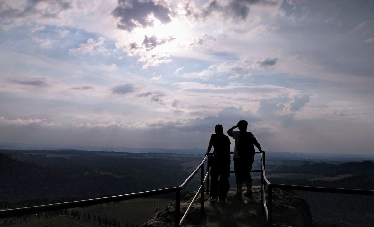 2 Menschen stehen auf einem Felsvorsprung mit Eisengeländer in der sächsischen Schweiz und schauen in die Ferne. Die Sonne scheint durch einen bewölkten Himmel.